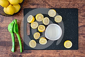 Fresh lemon juice in a white bowl, lemon halves on a black cutting board, wood table, basket of lemons, citrus squeezer