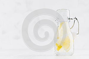 Fresh lemonade with lemon, slices, soda water in transparent yoke bottle on soft light white wood table, copy space.