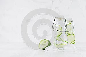 Fresh lemonades with green cucumber, soda water, ice cubes in transparent yoke bottles on soft light white wood table, copy space.