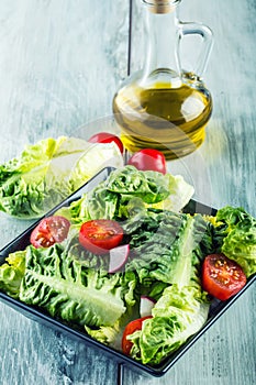Fresh lettuce salad with cherry tomatoes radish and carafe with olive oil on wooden table. Several ingredients of Mediterranean cu