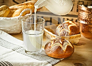 Fresh lye rolls in a wicker basket, process of pouring milk into a glass from pitcher, wood table, rustic style kitchen interior