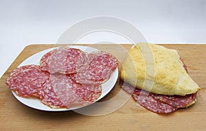 Fresh made salami sandwich, with slices of salami in a white dish, on wooden table and white background