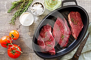 Fresh meat. Raw steak beef on a cast-iron grill frying pan, tomatoes, olive oil, spices and fresh rosemary on the kitchen table.