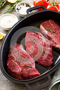 Fresh meat. Raw steak beef on a cast-iron grill frying pan, tomatoes, olive oil, spices and fresh rosemary on the kitchen table