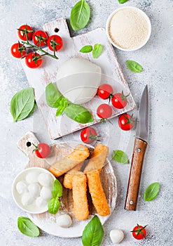 Fresh Mozzarella cheese on vintage chopping board with tomatoes and basil leaf and tray with cheese sticks on stone kitchen table