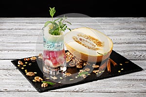 A fresh multi-colored alcoholic cocktail next to a melon and crushed walnuts on a wooden table background. Summer