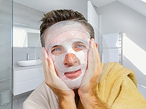 Young attractive and happy gay man in bathroom using beauty paper facial mask touching his face looking to the mirror smiling in