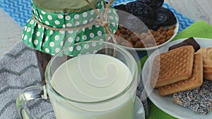 Fresh natural milk in a glass, a jar of jam, a white teaspoon, a saucer with dried fruits and a saucer with cookies and