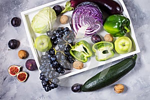 Fresh organic raw green and purple colored vegetables and fruits in white tray on dark stone background