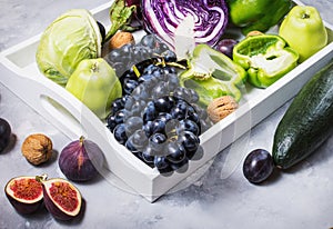 Fresh organic raw green and purple colored vegetables and fruits in white tray on dark stone background