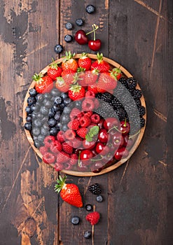 Fresh organic summer berries mix in round wooden tray on dark wooden table background. Raspberries, strawberries, blueberries,