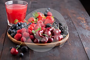 Fresh organic summer berries mix in round wooden tray with glass of juice on dark wooden table background. Raspberries,