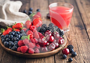 Fresh organic summer berries mix in round wooden tray with glass of juice on light wooden table background with linen towel.