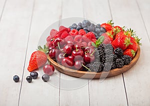 Fresh organic summer berries mix in round wooden tray on light wooden table background. Raspberries, strawberries, blueberries,