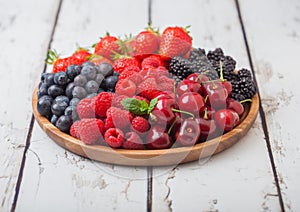 Fresh organic summer berries mix in round wooden tray on white wooden table background. Raspberries, strawberries, blueberries,