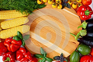 Fresh organic vegetables and herbs around wooden board: bell pepper, corncobs, avocado, eggplants, cherry tomatoes, basil, dill