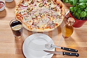 Fresh pizza with ham, hot pepper and capers and black olives on wooden table in restaurant.