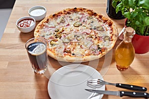 Fresh pizza with ham, hot pepper and capers and black olives on wooden table in restaurant.