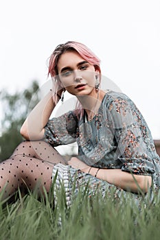 Fresh portrait fashionable young woman model with chic pink hair in stylish blue dress with floral print on grass in field on