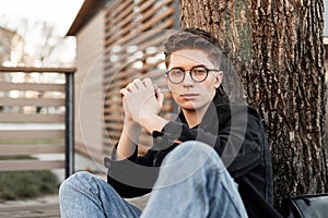 Fresh portrait serious young hipster man in fashionable glasses in denim jacket in blue jeans with hairstyle on street. Attractive