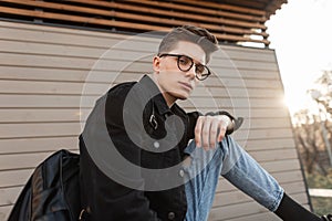 Fresh portrait serious young hipster man with vintage glasses in denim jacket with backpack on street at sunset. Attractive modern