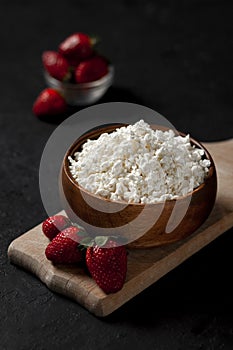 Fresh pure homemade cottage cheese in a wooden plate with strawberries on a black table