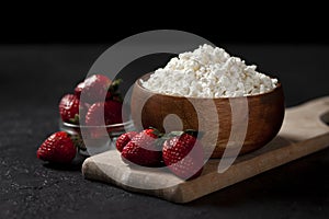 Fresh pure homemade cottage cheese in a wooden plate with strawberries on a black table, healthy food against a dark