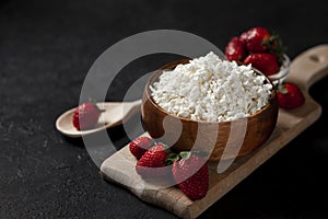 Fresh pure homemade cottage cheese in a wooden plate with strawberries on a black table, healthy food against a dark