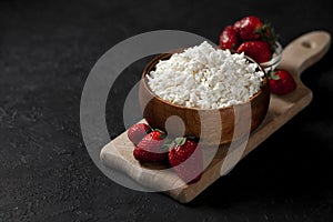 Fresh pure homemade cottage cheese in a wooden plate with strawberries on a black table, healthy food against a dark background