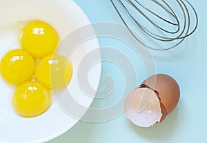 Fresh raw chicken yolks with whisk, eggs shells on the blue background. Baking or food preparation concept