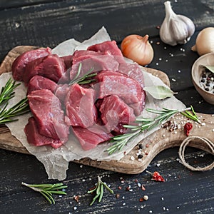 Fresh raw chopped beef on an olive board, spices, herbs and vegetables on dark wooden background.