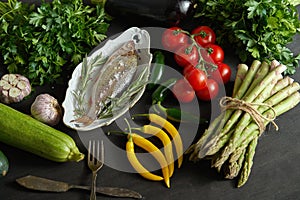 Fresh raw dorada fish in a white dish with a set of vegetables on a black table