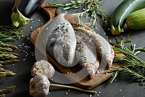 Fresh raw dorada fish on a wooden board with a set of vegetables on a black table