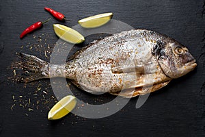 Fresh raw dorado on black slate stone board with spices, herbs, lemon and red hot pepper. Sea bream fish ready for cooking. Top