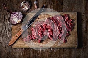 Fresh raw meat cut into pieces in the process of cooking beef stroganoff lies on a wooden cutting board with a kitchen knife,