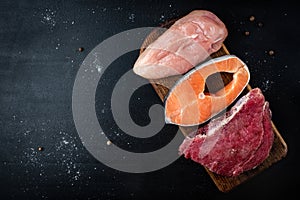 Fresh raw meat, salmon and chicken fillet on wooden cutting board on black background. Natural food high in protein. Top view.