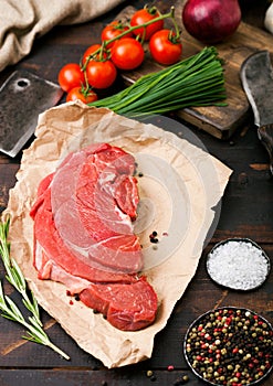 Fresh raw organic slice of braising steak fillet on butchers paper with fork and knife on dark wooden background. Red onion,