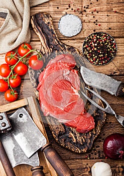 Fresh raw organic slice of braising steak fillet on chopping board with fork and knife on wooden background. Red onion, tomatoes