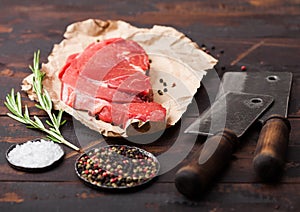 Fresh raw organic slice of braising steak fillet on paper with meat hatchets on dark wooden background. Salt and Pepper