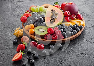 Fresh raw organic summer berries and exotic fruits in round wooden plate on black kitchen background. Papaya, grapes, nectarine,