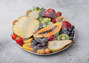 Fresh raw organic summer berries and exotic fruits in white plate on light background. Pineapple, papaya, grapes, nectarine,