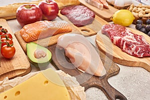 Fresh raw poultry, meat, fish on wooden cutting boards near apples, lemon, cheese, cherry tomatoes and half of avocadoes