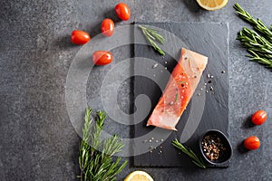 Fresh raw salmon fillet on black stone board with salt, peppers, lemon, and rosemary on dark background, top view, copy space.