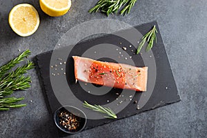 Fresh raw salmon fillet on black stone board with salt, peppers, lemon, and rosemary on dark background, top view. Restaurant menu