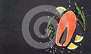 Fresh raw salmon fish steak with cooking ingredients, herbs and lemon on black background, top view