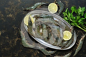Fresh raw tiger shrimps on a metal plate with ice, lemon and parsley on a dark background. Top view, copy space
