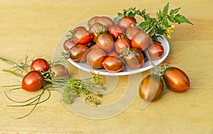 The fresh red and green bright tomatoes cherry and green tomato leaves with yellow flowers lie on a white plate on a yellow wooden