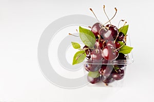 Fresh red juicy cherries with green leaves and tails in transparent plastic box on soft light white wood board, copy space.