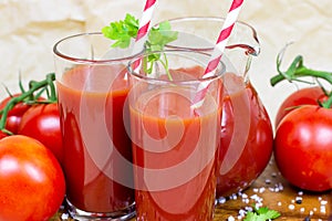 Fresh red tomato juice in a glass with a straw and jar with tomatoes on light wooden background.