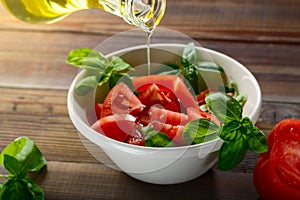 Fresh red tomatoes are sliced in a white ceramic plate with basil herb and olive oil on a wooden table